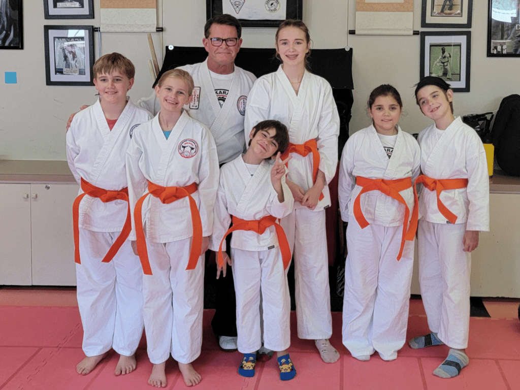 Martial arts students, wearing white uniforms with orange belts, pose with an instructor in a dojo decorated with martial arts memorabilia.